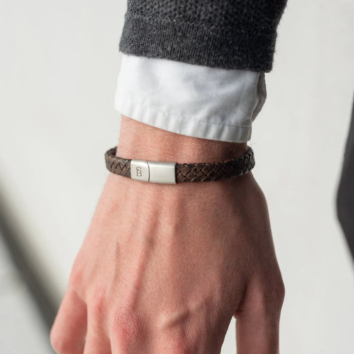 A close-up image of a person's wrist wearing a Steel and Barnett Riley - Vintage Brown bracelet with a branded metal clasp. The person is dressed in a white shirt layered with a dark-colored sweater. The background is blurred and neutral.