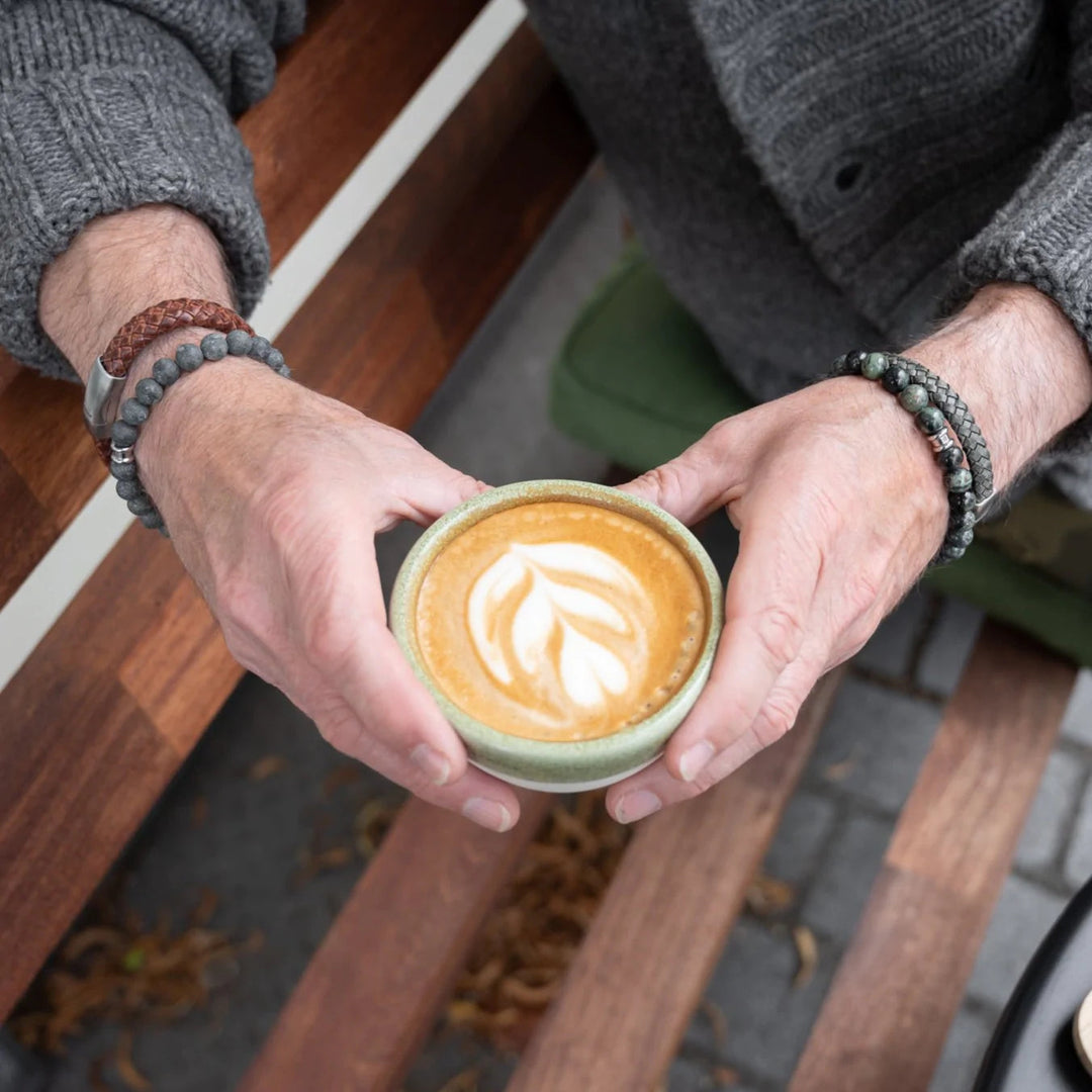 A person with Riley - Dark Green by Steel and Barnett stainless steel and braided leather bracelets on both wrists holds a cup of latte with a leaf design in the foam. The person is wearing a gray sweater and sitting on a wooden bench, adorned with their bracelets' branded metal clasps. Fallen leaves are scattered on the ground below.