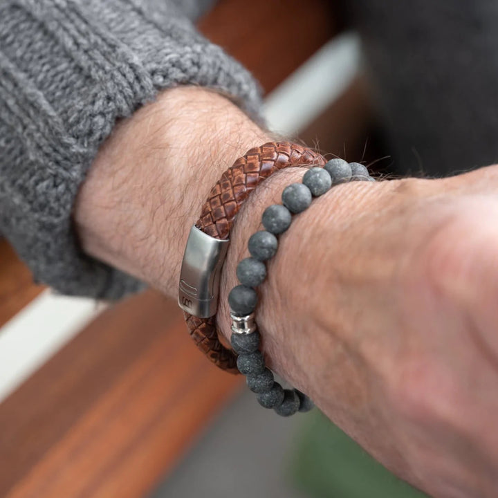 Close-up of a person's wrist wearing two bracelets: a genuine braided leather bracelet with a stainless steel clasp and a beaded gray stone bracelet. The person is also wearing Steel and Barnett's Cornall - Caramel bracelet and a gray knitted sweater, showcasing modern minimalism.