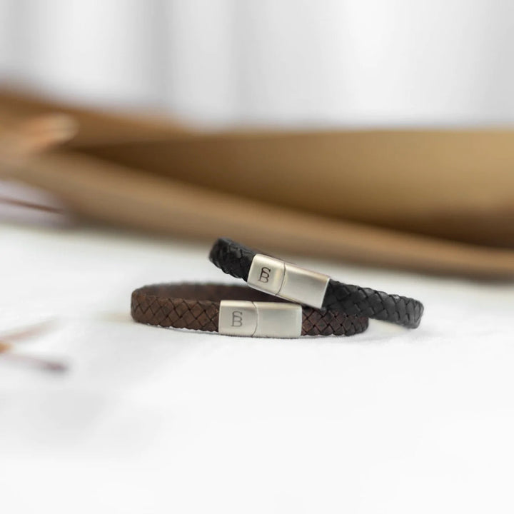 Two sleek leather bracelets lie side by side on a white surface. One is dark brown, and the other is black. Both feature a genuine braided leather design and have polished branded metal clasps engraved with a "B." A blurred background suggests a cozy indoor setting. The dark brown bracelet is the Riley - Vintage Brown by Steel and Barnett.