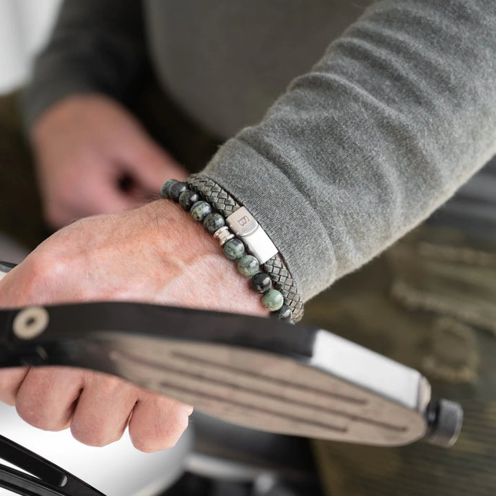 A person wearing a gray long-sleeve shirt is shown gripping a bicycle handlebar. They sport green-beaded bracelets and a braided leather bracelet on their wrist, featuring a branded metal clasp from Steel and Barnett. The focus is on the Riley - Dark Green bracelets and the hand, with a blurred background.