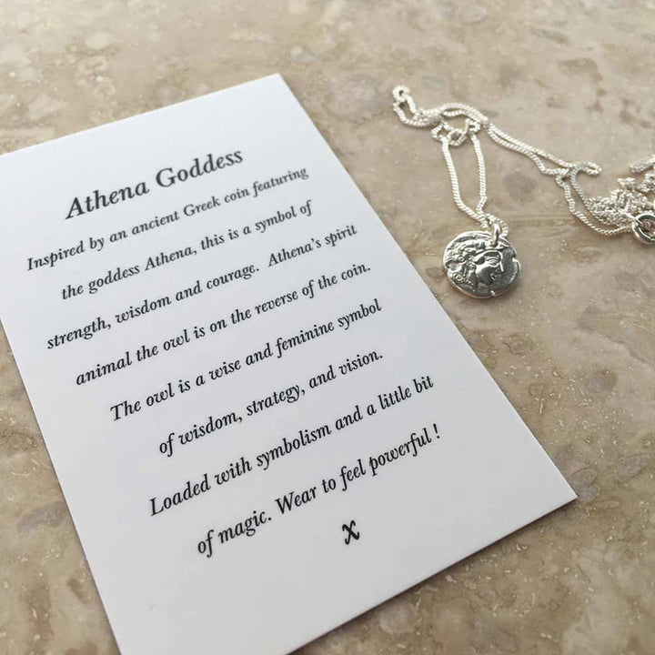 An Athena Coin Necklace Silver by Andrea Mears Jewellery featuring a pendant of the Greek goddess Athena rests on a marble surface, alongside an information card. The card explains Athena's symbolism of strength, wisdom, and courage and highlights the owl on the coin as a symbol of wisdom and strategy.