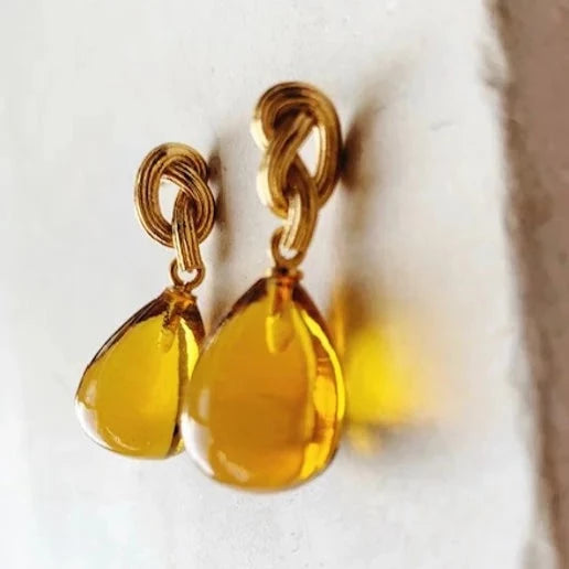 The Shyla Synthea Earrings feature teardrop-shaped yellow pendants hanging from textured gold loops, shown against a light background.