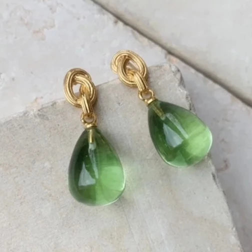 The Shyla Synthea Earrings in green feature elegant gold knots and drop gemstones, displayed on a light stone surface—hypoallergenic for stylish everyday wear.