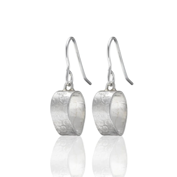 A pair of elegant sterling silver Talún Drops by Lynsey de Burca with a curved, textured surface. Each earring is suspended from a simple hook. The surface appears to have a hammered or brushed finish, giving it a unique, handmade jewellery look. The overall design is minimalistic yet sophisticated.