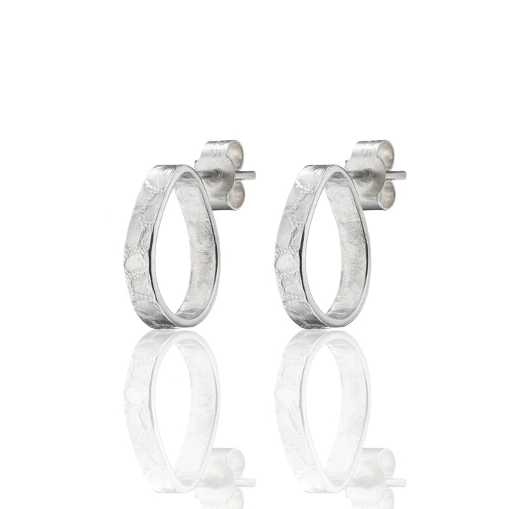 A pair of sterling silver, textured raindrop stud earrings with stud backs. The Talún Studs by Lynsey de Burca are shown standing upright, with their reflections visible below them on a white background.