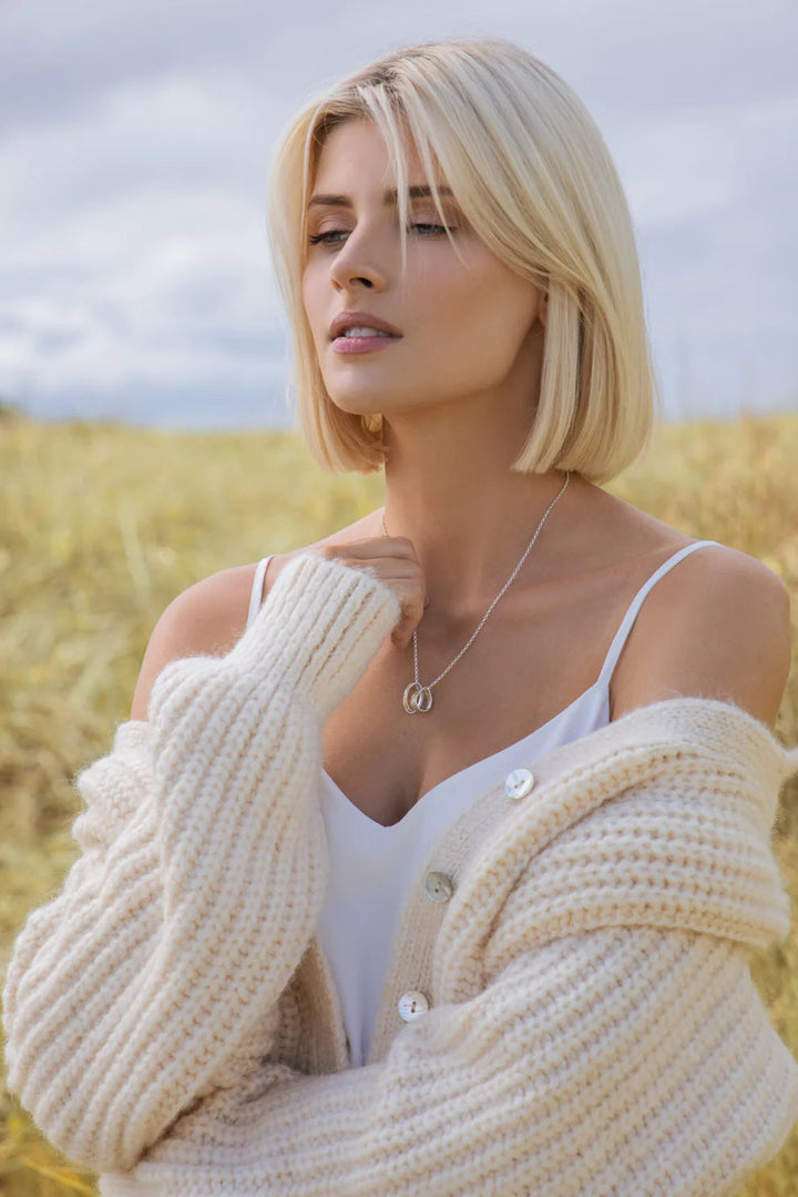 A person with blond hair is standing in front of a field, wearing a white camisole and a cozy cream-colored knitted cardigan. Adorned with the Talún Triple Neckpiece by Lynsey de Burca, they appear serene, gazing slightly to the side with a neutral expression.