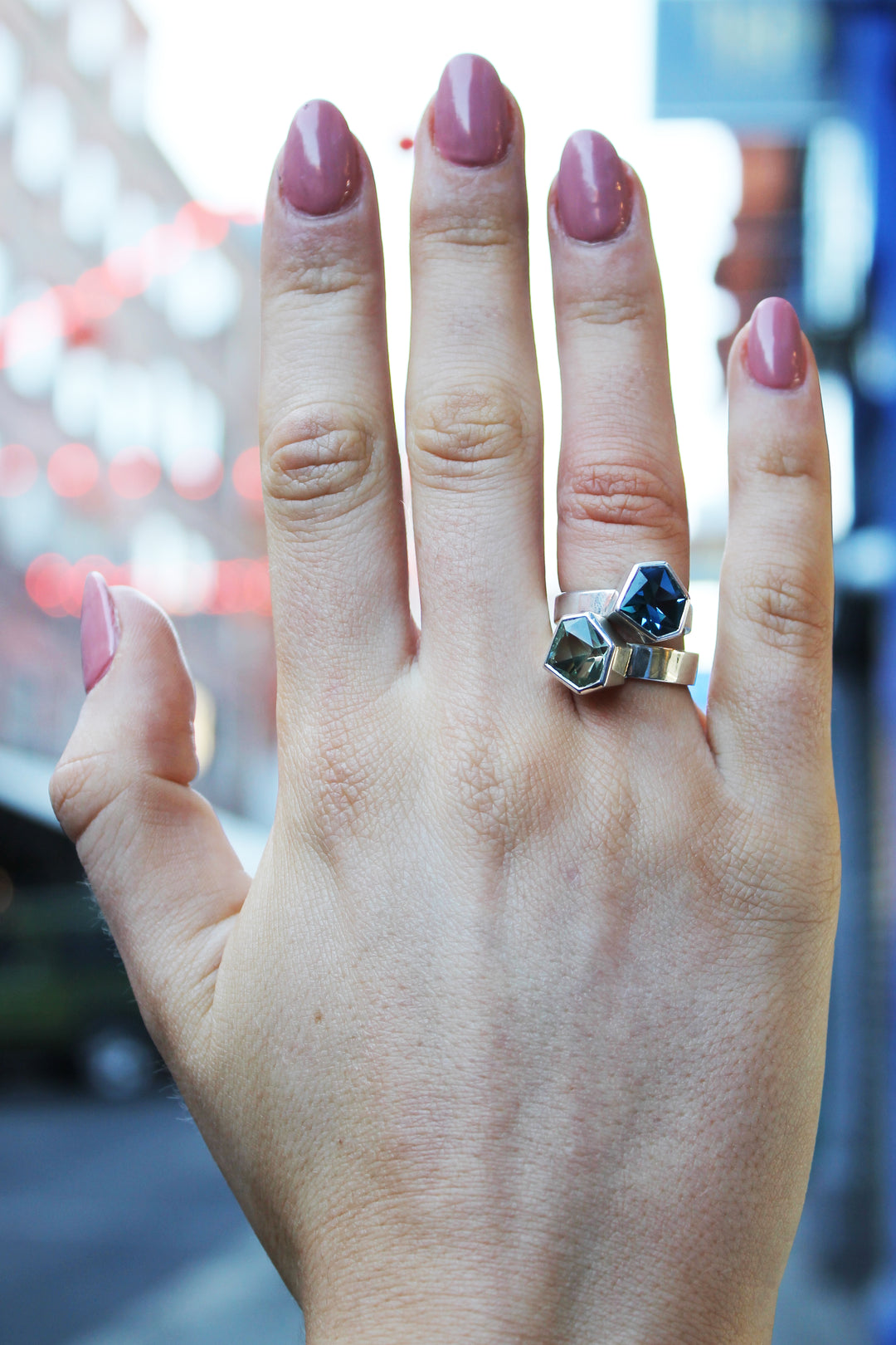 two rings, the first one, closer to the hand is a Silver ring with large, irregular hexagonal green amethyst in a silver bezel. Band is flat and 5mm wide.The second is a Silver ring with large, irregular hexagonal blue topaz in a silver bezel. Band is flat and 4mm wide