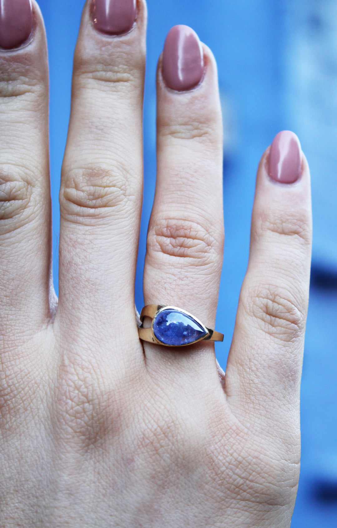 9ct yellow gold ring with teardrop shaped cabochon Tanzanite, which is a dark blue purple stone, bezel set in 9ct yellow gold. Irregular shaped band, tapers from a thicker width with cut-out detail and tapers down to a thinner band. Shown here on a ring finger