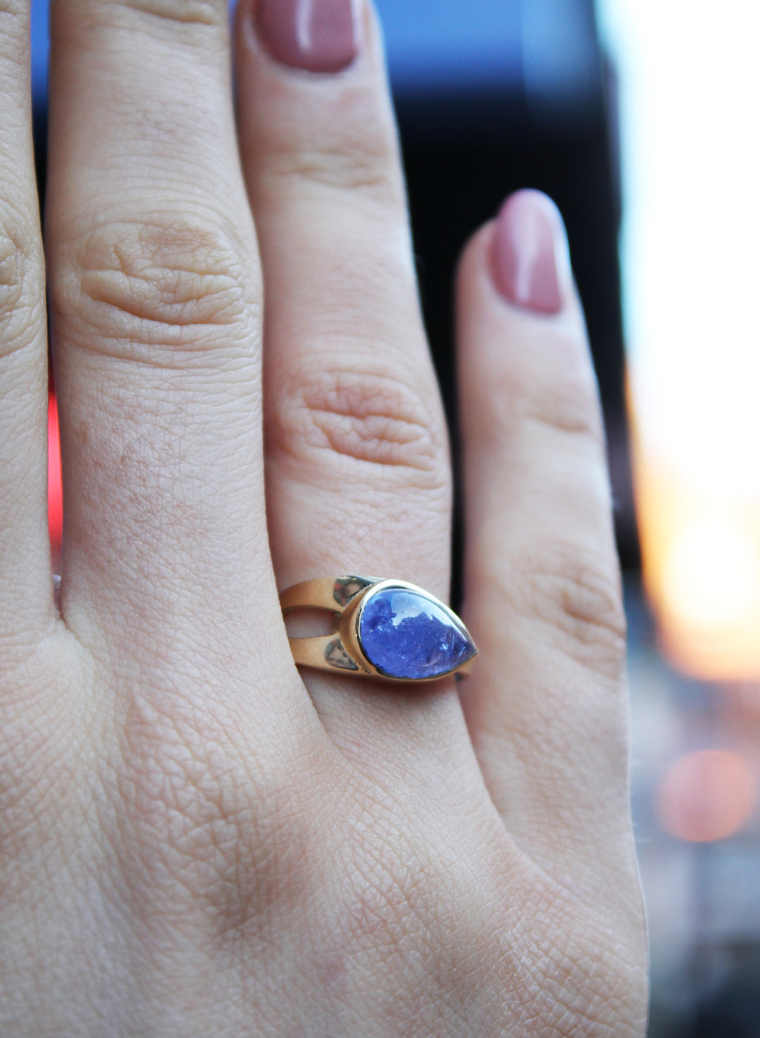 9ct yellow gold ring with teardrop shaped cabochon Tanzanite, which is a dark blue purple stone, bezel set in 9ct yellow gold. Irregular shaped band, tapers from a thicker width with cut-out detail and tapers down to a thinner band. Shown here on a ring finger