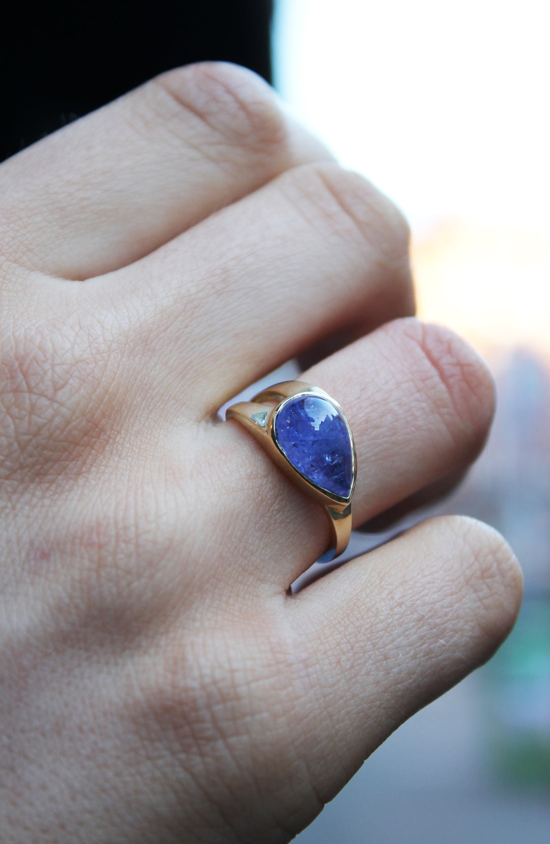 9ct yellow gold ring with teardrop shaped cabochon Tanzanite, which is a dark blue purple stone, bezel set in 9ct yellow gold. Irregular shaped band, tapers from a thicker width with cut-out detail and tapers down to a thinner band. Shown here on a ring finger