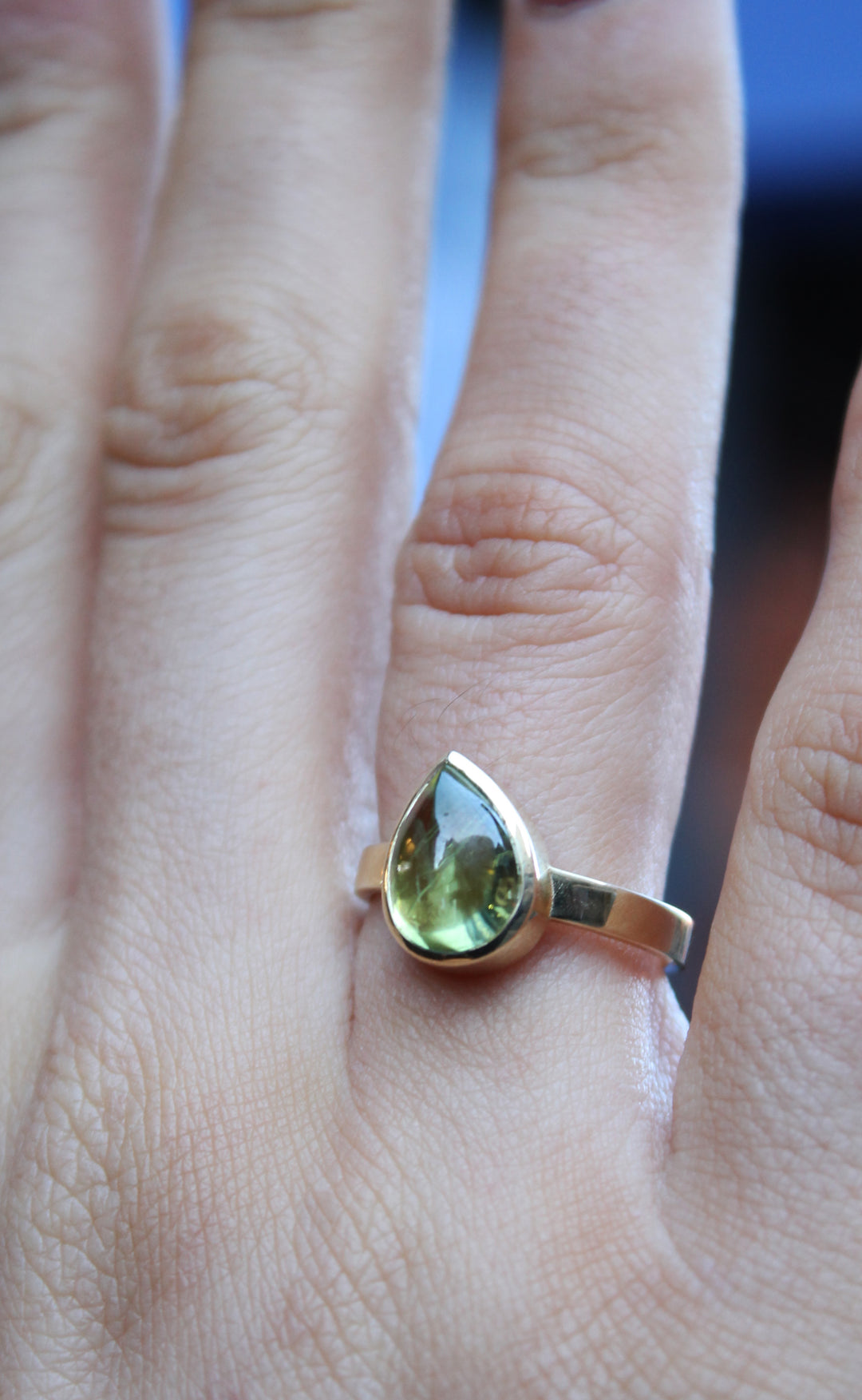 9ct yellow gold ring with a teardrop shaped peridot cabochon, which is a moss green gem, in a bezel setting. The band is flat in shape and is 3mm wide. The 9ct yellow gold band is 3mm wide. Shown here on a ring finger.