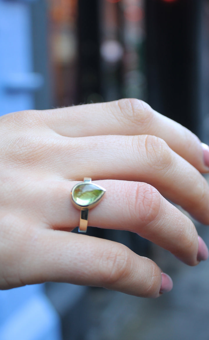 9ct yellow gold ring with a teardrop shaped peridot cabochon, which is a moss green gem, in a bezel setting. The band is flat in shape and is 3mm wide. The 9ct yellow gold band is 3mm wide. Shown here on a ring finger