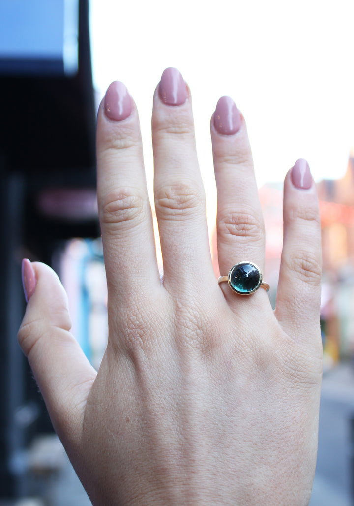  9ct yellow gold ring with a round dark green tourmaline cabochon in a bezel setting. The 9ct yellow gold band is D shaped widest at the back at 3mm wide. Shown here on a ring finger.