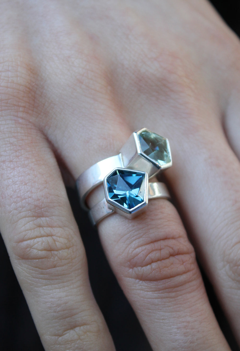 two rings, the first one, closer to the hand is a Silver ring with large, irregular hexagonal green amethyst in a silver bezel. Band is flat and 5mm wide.The second is a Silver ring with large, irregular hexagonal blue topaz in a silver bezel. Band is flat and 4mm wide