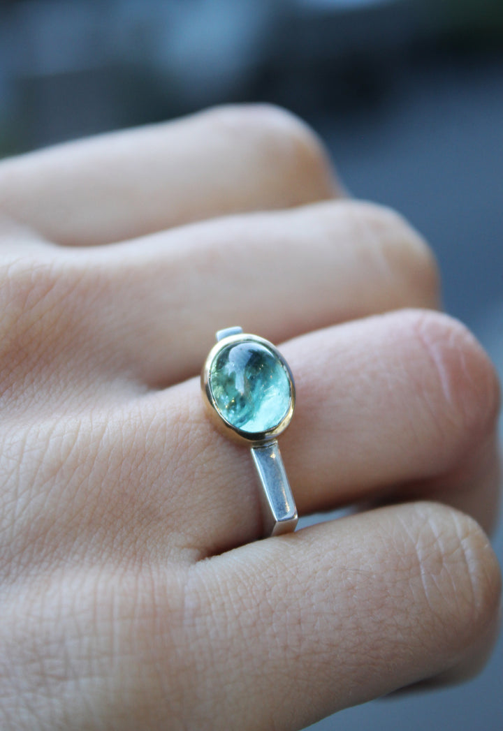 Silver ring with green tourmaline in 9ct yellow gold bezel . Tourmaline is off centre. The top of the ring is rectangular making the band D shaped. Shown here on a ring finger.