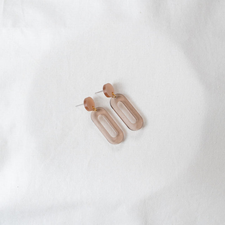 A pair of Taupe Oval Acrylic Earrings by Studio Nok Nok lying on a plain white fabric. The earrings feature a geometric design with a small circular stud connected to an elongated, rounded rectangle shape. Made from recycled acrylic, the material appears smooth with a matte finish in a neutral color.