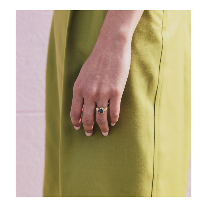 A close-up of a person's hand wearing a gold ring with a dark triangular stone, set against a solid olive green dress.