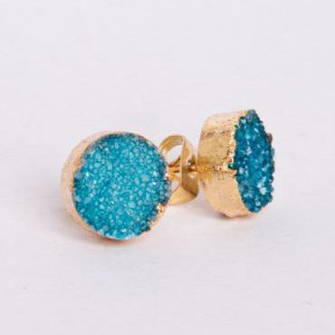 A pair of Teal Blue Druzy Set In Gold Stud Earrings by Watermelon Tropical featuring a textured, teal blue druzy center with gold-toned edges. The earrings have a slightly rough, crystalline appearance and are set against a plain white background.