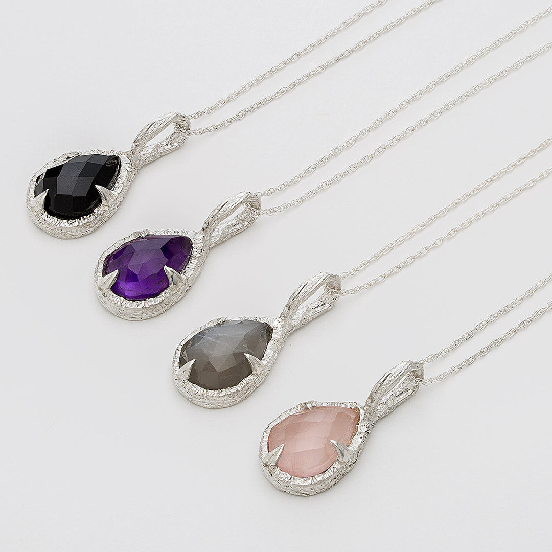 Four elegant Thorn Teardrop Pendants on sterling silver chains from Abbie Dixon, each featuring a unique teardrop-shaped gemstone: black, purple, grey, and light pink. The gemstones are encased in textured silver settings, highlighting their distinct colors and facets. One pendant showcases a shimmering moonstone for added allure.