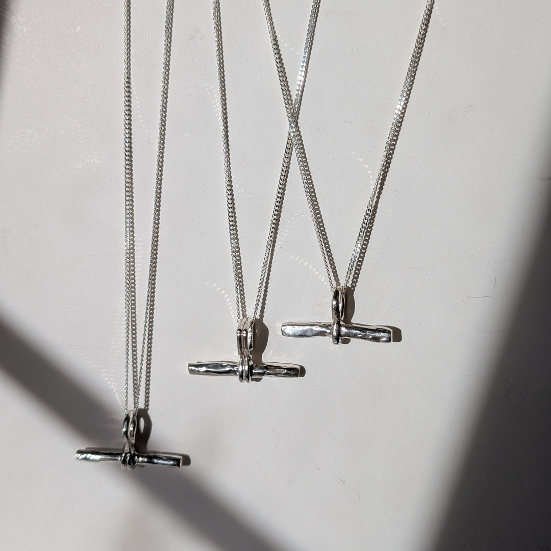 Three Becca Calmont T-Bar Pendant necklaces in sterling silver are arranged on a white surface, with sunlight casting soft shadows of the simple horizontal pendants and their curb chains.