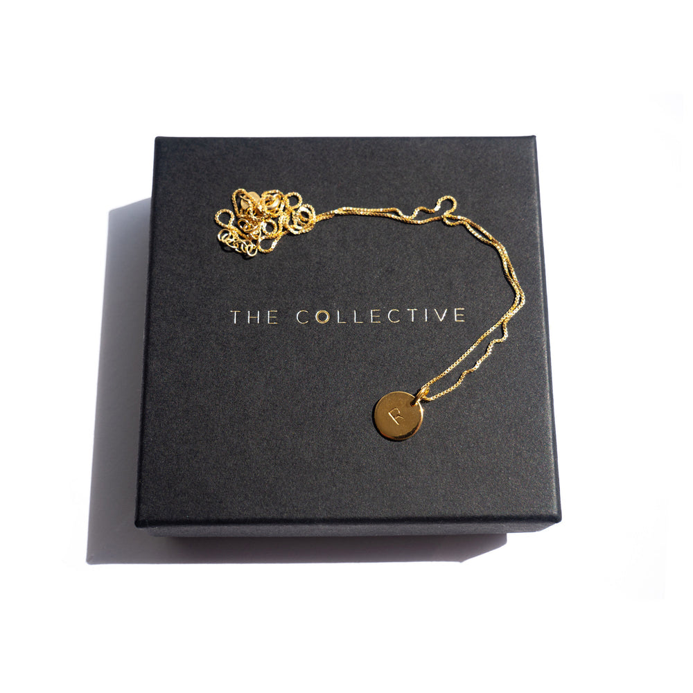 A delicate gold necklace featuring a circular pendant engraved with the letter "R" is displayed on a black box. The box, adorned with the words "Personalised Charms" in gold letters, showcases the partially coiled necklace at the top left corner. The hand stamped charm adds a personal touch to this elegant piece of Personalised Small Disc Charm in Gold.