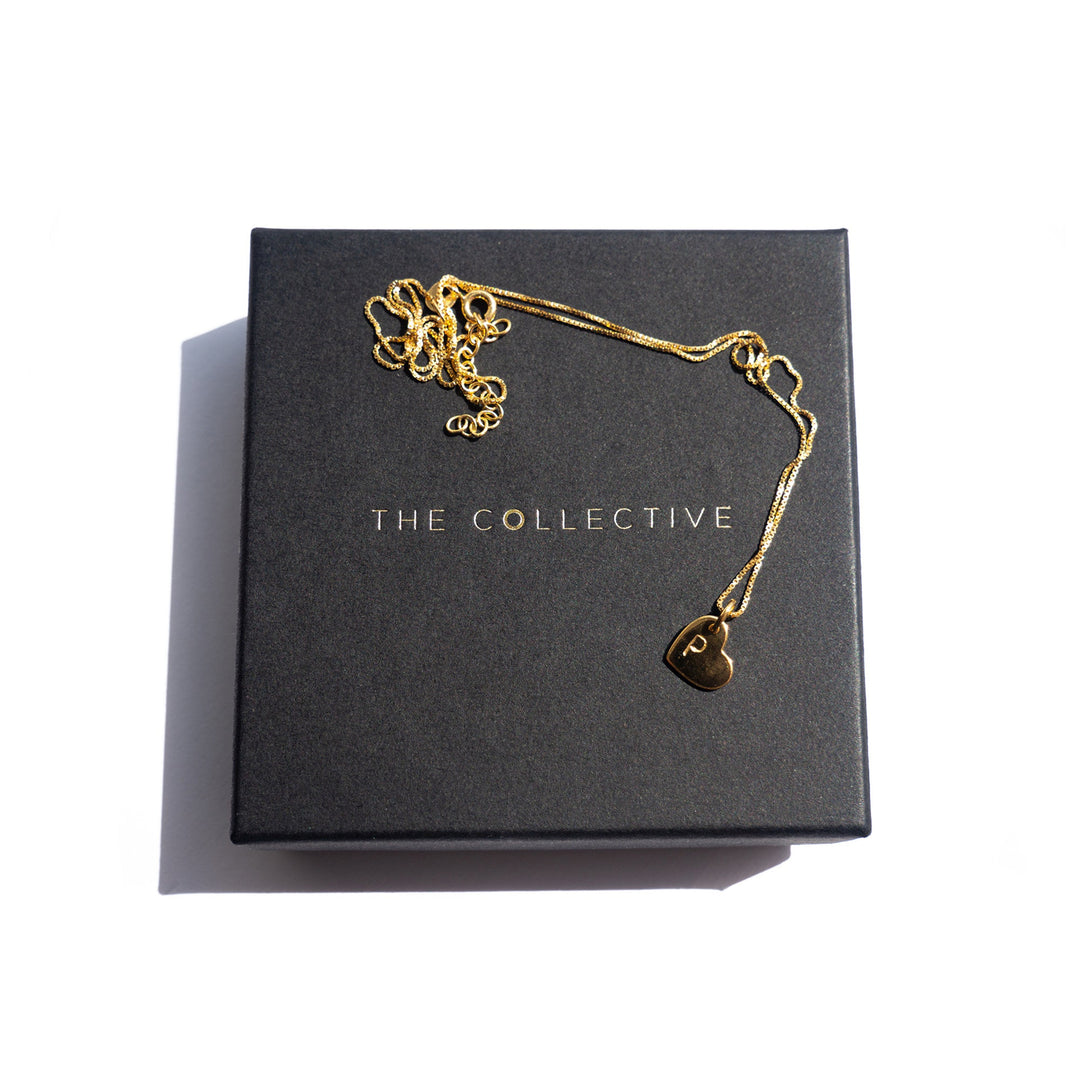 A delicate gold plated silver necklace with a small hand stamped charm, engraved with the letter 'P', is resting on a black box. The box has "PERSONALISED CHARMS" written in silver capital letters on its surface. The adjustable oval belcher chain is loosely arranged on the box.