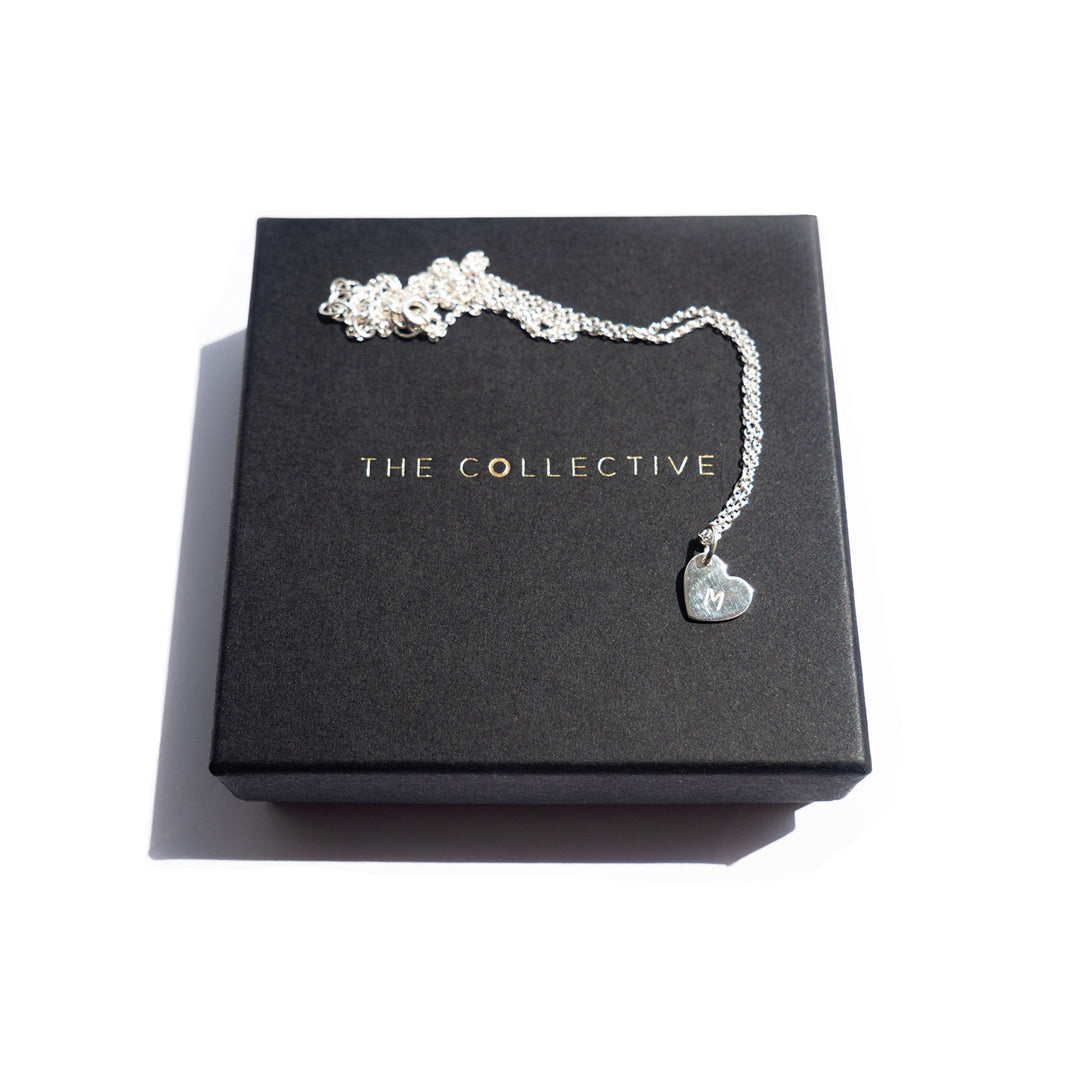 A **Personalised Small Heart Charm in Silver** from **Personalised Charms** is displayed on a black box with gold lettering that reads "THE COLLECTIVE." The oval belcher chain is delicately coiled, highlighting the pendant and its lobster claw fastening. The background is white.