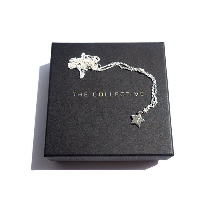 A Personalised Small Star Charm in Silver by Personalised Charms is displayed on a black box. The box has the text "THE COLLECTIVE" written in gold letters. The hand-stamped, oval belcher chain and star pendant are neatly arranged with a white background casting a light shadow.