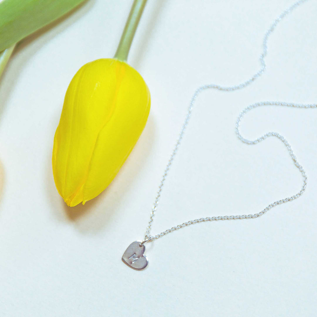 A vibrant yellow tulip lays next to a delicate silver Personalised Small Heart Charm in Silver with an oval belcher chain and lobster claw fastening on a white background. The necklace, from Personalised Charms, features simple silver charms, and the scene exudes elegance and freshness.