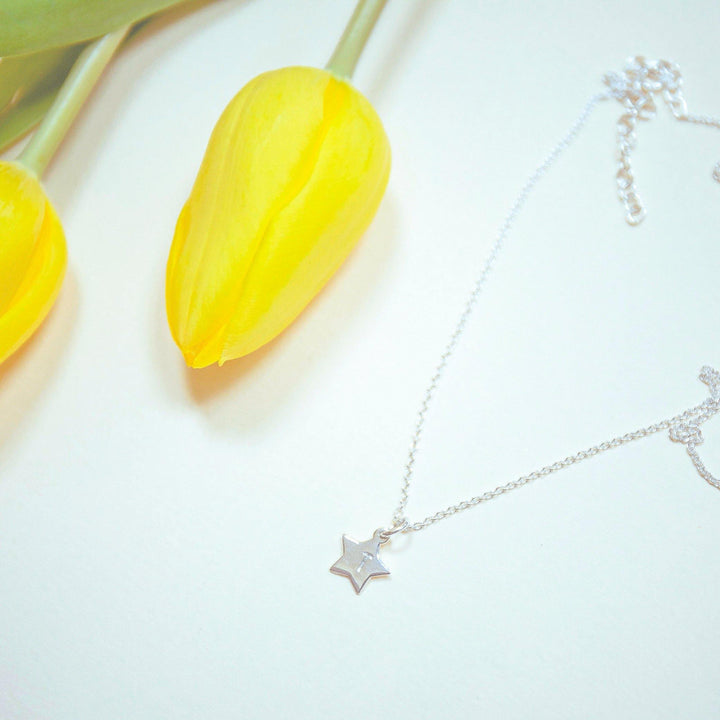 A delicate necklace with a star-shaped pendant lies on a light surface beside two vibrant yellow tulips. The oval belcher chain is coiled, and the scene is gently lit, emphasizing the soft, fresh appearance of the tulips and the subtle shine of the hand-stamped Personalised Small Star Charm in Silver by Personalised Charms.