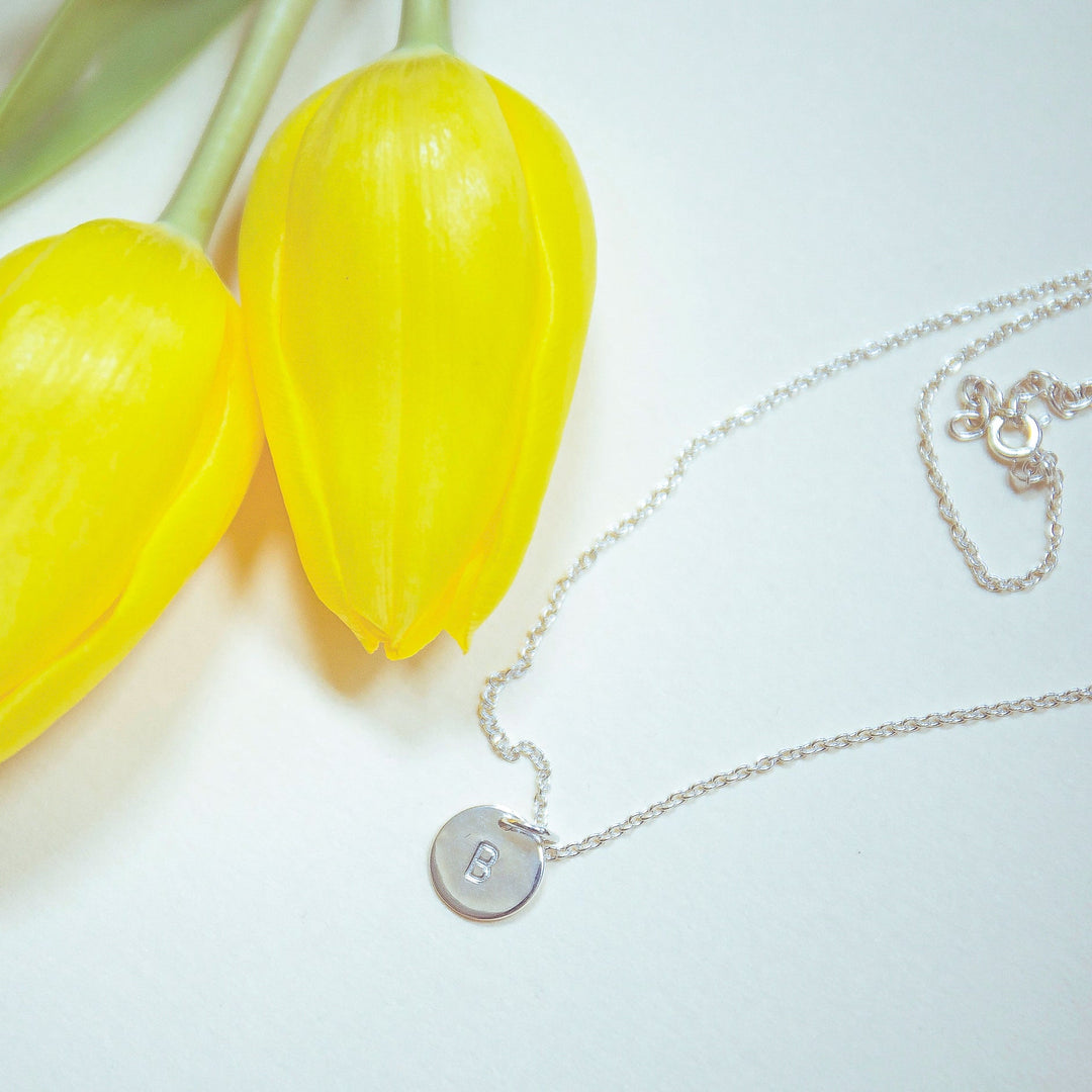 A close-up image shows two bright yellow tulips and a delicate silver necklace. The Personalised Charms' Personalised Small Disc Charm in Silver, with an oval belcher chain, features a round pendant with the letter "B" hand stamped on it. The items are placed on a smooth, white surface.