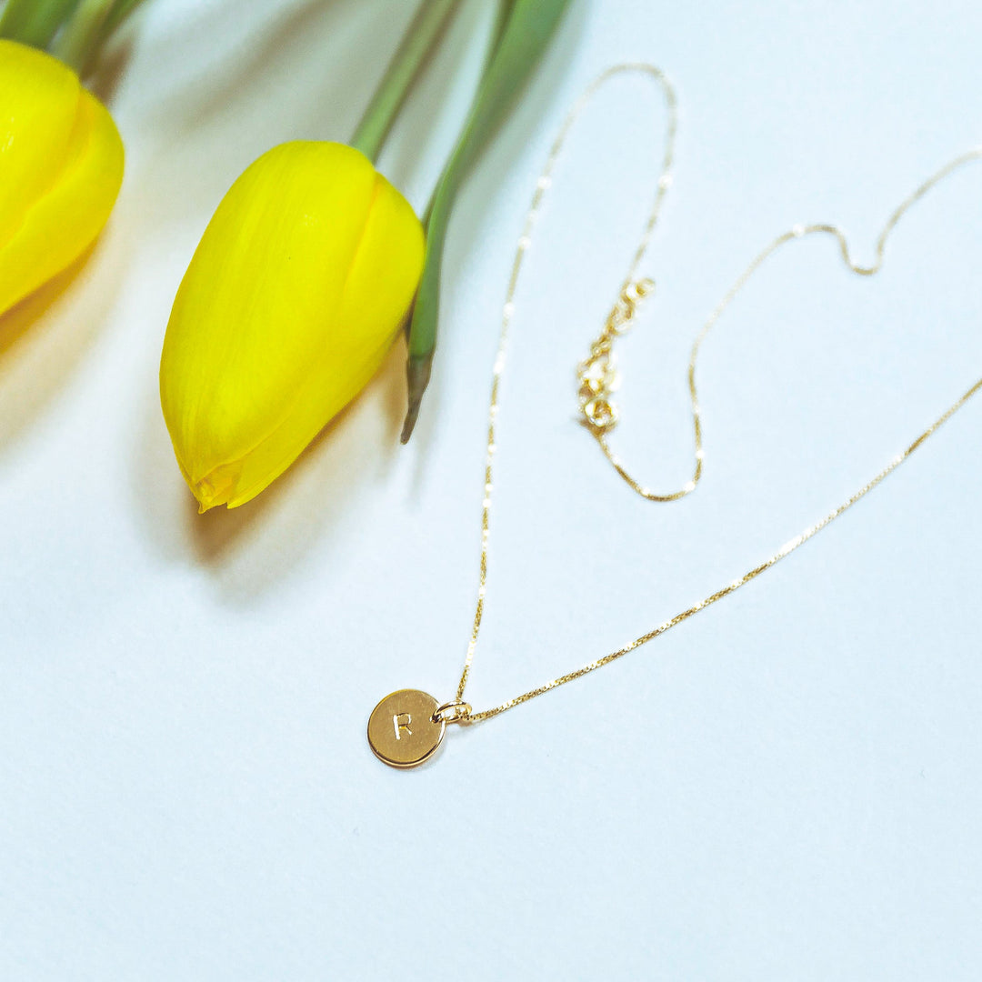 A delicate Personalised Small Disc Charm in Gold by Personalised Charms with a round pendant hand stamped with the letter "R" is laid on a white surface. Next to the necklace, there are two vibrant yellow tulips, accentuating the elegance of the scene.