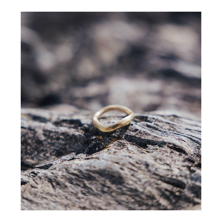 A thin, wavy gold ring resting on a textured, weathered piece of wood with a blurred natural background.