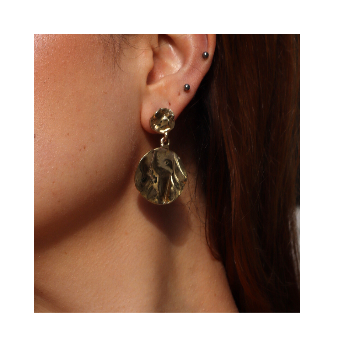A close-up of a woman's ear showcases three small studs on the upper ear and the Tide Drop Earrings—a large, gold, wavy, round dangle by Madeleine Sculptural Jewellery—on the earlobe. Brown hair is partially visible in the background.