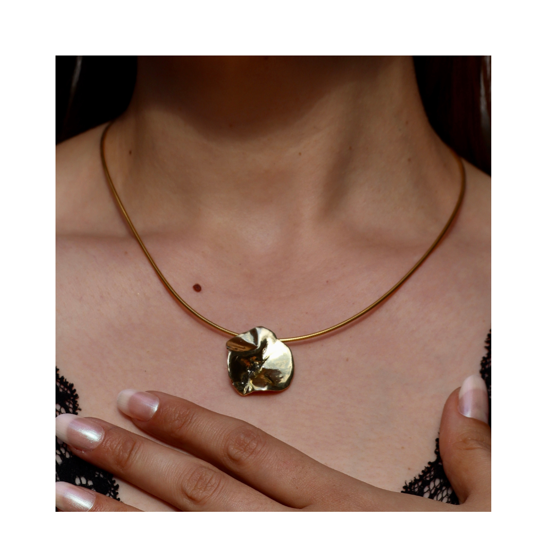 A woman wears the Tide Pendant by Madeleine Sculptural Jewellery—a gold necklace with a large, textured, leaf-shaped pendant featuring organic lines. Her hand touches her chest over a black lace top.
