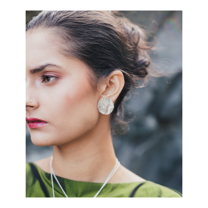 A close-up side profile of a woman wearing a round, textured light gray stone earring, with her hair tied back and a green top visible against a blurred dark gray background.