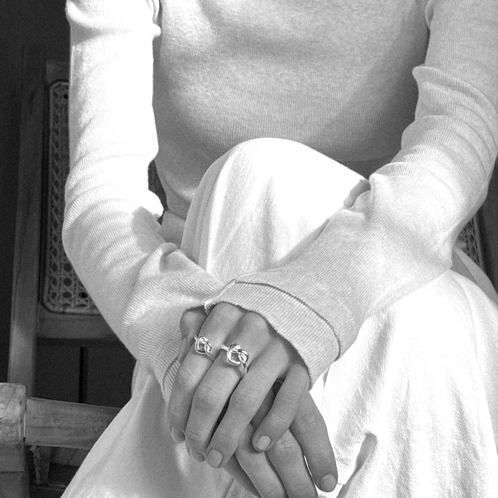 A person sits with hands on their knee, wearing two chunky rings including the Jane Koenig Tied Heart Ring in sterling silver. They wear a long-sleeved, light top and skirt. The black-and-white image highlights the torso and hands.