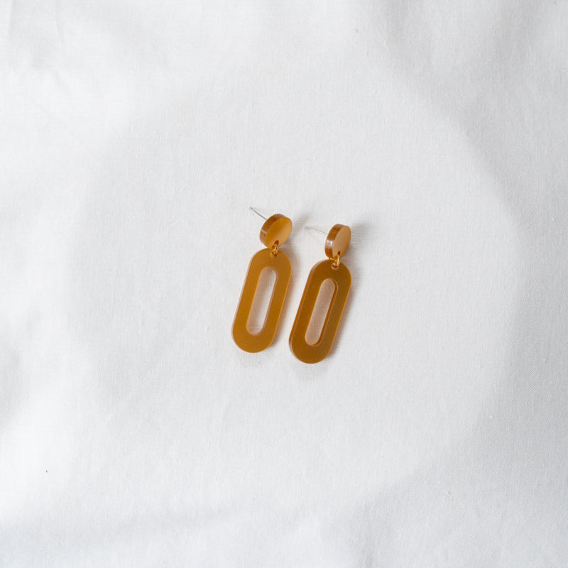 A pair of perfect yellow-orange earrings with a modern, geometric design. Each earring consists of an oval-shaped loop connected to a hollowed-out oblong piece, crafted from recycled materials. The Timeless 04 Oval Acrylic Earrings by Studio Nok Nok are lying on a white fabric background, epitomizing the beauty of handmade jewelry.