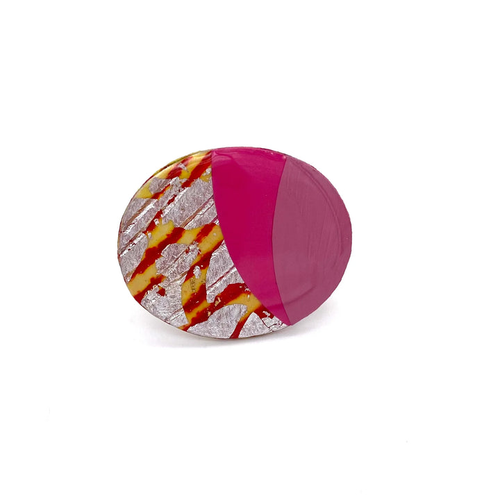 A round, handmade modern art sculpture with one half painted in solid magenta and the other half featuring a textured pattern of silver and yellow with red striped accents is displayed against a plain white background. The product is Tonn Ring in Orange/Silver/Rose/Hot Pink by Rothlú.