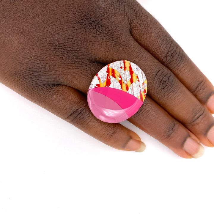 A close-up of a hand with dark skin wearing a large, oval statement ring. The Rothlú Tonn Ring in Orange/Silver/Rose/Hot Pink features an abstract design with sections of pink, red, and reflective silver patterns, creating a bold, artistic look. The hand is positioned against a white background.