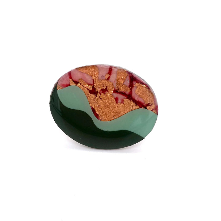 An oval-shaped abstract stone features green, brown, and red hues, with a textured design that includes streaks and patches resembling a landscape. The stone has a glossy finish and appears to be handcrafted, set in hypoallergenic silver-plated metal as the exquisite Tonn Ring in Red/Rose Gold/Mint/Olive by Rothlú.