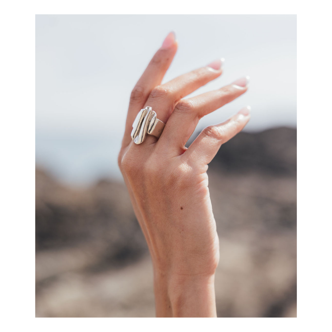 A close-up of a hand wearing a wide, sculptural silver ring with layered, organic shapes, set against a blurred outdoor desert background.