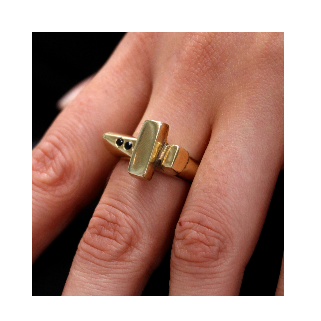 A close-up of a hand wearing Madeleine Sculptural Jewellery's Totem Ring in 9K Gold, featuring a geometric rectangular bar and small black stones in a pointed design—an elegant piece of handmade jewellery.
