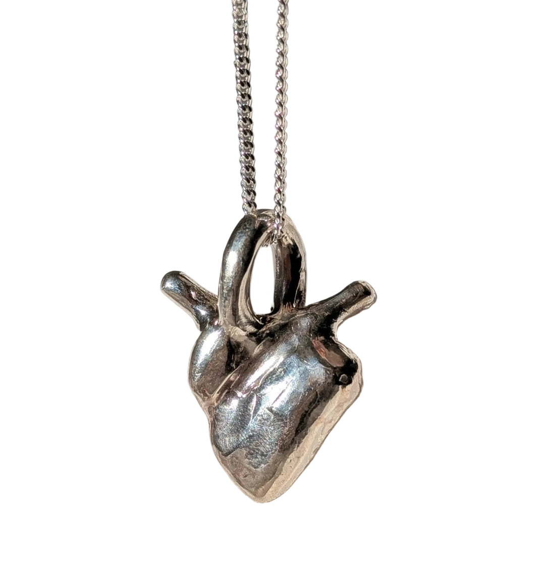 The True Love Heart Pendant by Becca Calmont, crafted from recycled sterling silver, features a silver anatomical heart on a thin chain necklace, displayed on textured white fabric with soft natural light and gentle shadows.