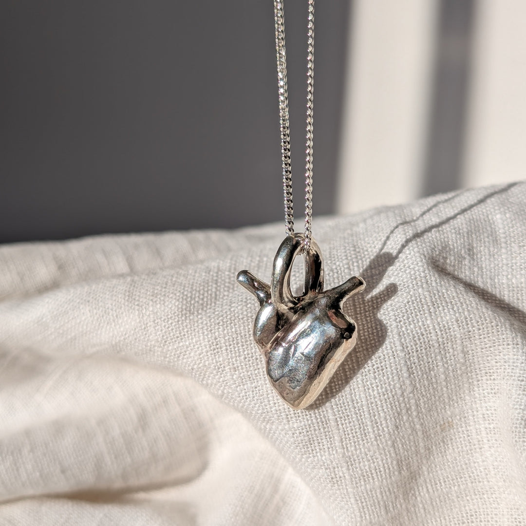 Silver anatomical heart necklace on a chain, resting on a textured surface with a neutral background.