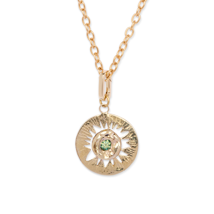 Introducing the Tundra Necklace by Madeleine Blaine—a statement piece inspired by the beauty of polar landscapes, featuring a round textured gold pendant with a radiant sunburst design and a vibrant green gemstone at its center, elegantly suspended from a gold chain with elongated oval links.
