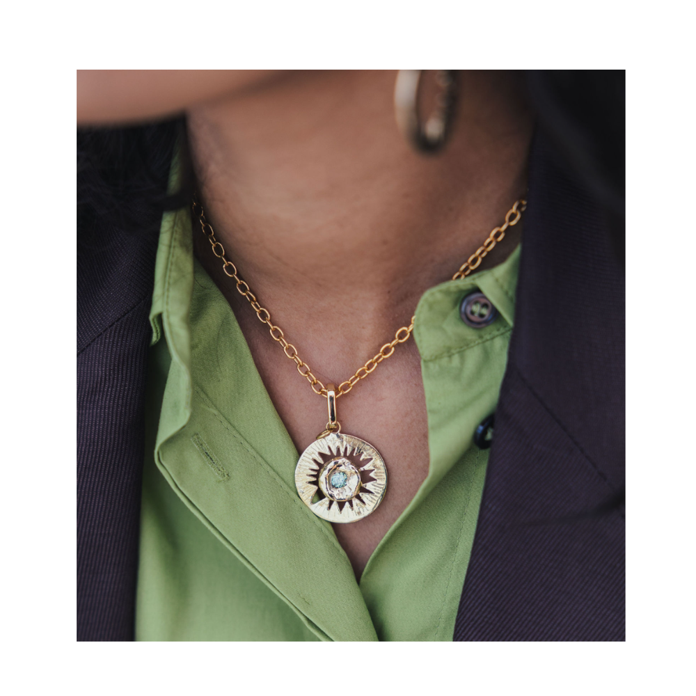 A close-up shows a person wearing the Madeleine Blaine Tundra Necklace—a gold chain accented with a sunburst pendant—layered over a green shirt and dark blazer. This striking statement piece, with only part of the face and torso in view, instantly draws the eye.