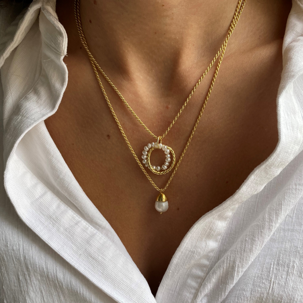 Close-up of a person wearing two gold necklaces. One features a circular pendant adorned with pearl-like beads, showcasing elegant 18k gold plating, while the other has a single pearl-like drop pendant from Bonvo's Twisted Pearl Pendant Rope Chain collection. The person is dressed in a white, slightly unbuttoned shirt.