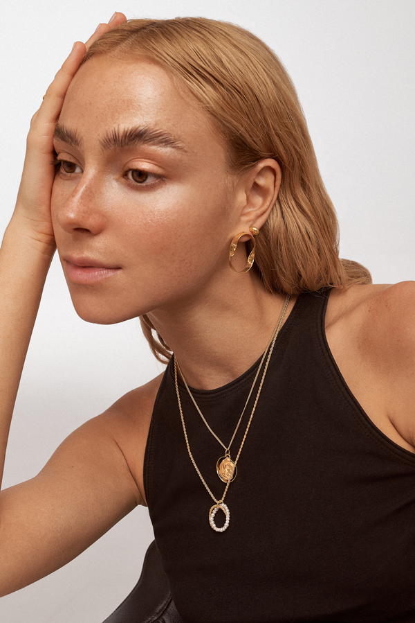 A person wearing a sleeveless black top poses with their hand resting on the side of their head. They have long light hair and are adorned with multiple gold necklaces, including a Twisted Pearl Pendant Rope Chain by Bonvo, and a large gold earring. The background is plain off-white.